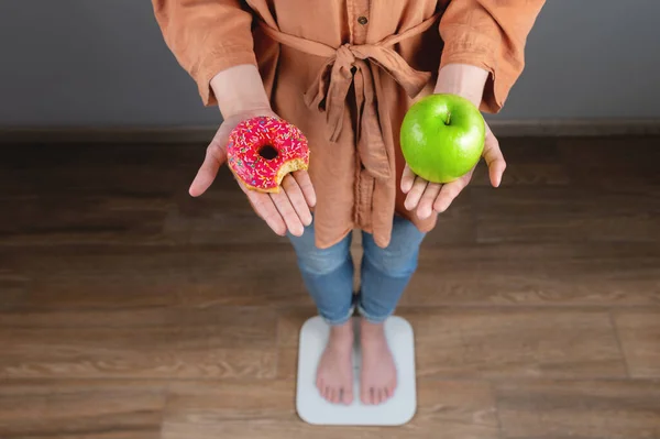 woman holding apple and donuts and standing on scales at home, health concept. unrecognizable girl weighs herself and makes a choice in favor of a healthy or unhealthy diet.