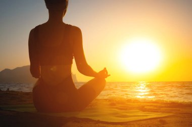 Kız yoga yapıyor ve plajda lotus pozisyonunda meditasyon yapıyor. Şafak vakti deniz kenarında genç bir kadının silueti, sağlıklı yaşam tarzı konsepti, arkadan görüş..