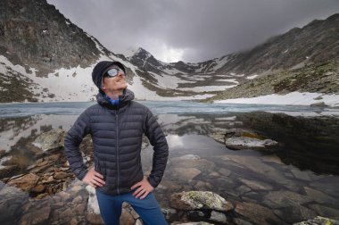 Genç bir turist dağın tepesinde bir dağ gölünün arka planına karşı duruyor. Kameraya gülümseyen mutlu adam. Turizm, spor ve seyahat konsepti