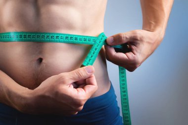 A muscular athlete poses shirtless and measures his waistline with a measuring tape. Close-up of an athlete measuring his body volume and the result of diet and training clipart