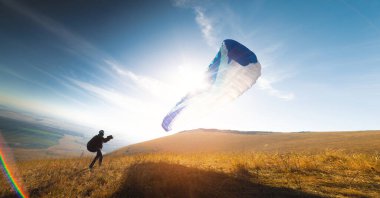 Mavi paraşütlü bir paraglider havalanıyor. Bir adam havalanıyor ve sarı bir tarlaya iniyor. Bir adam havada açık bir paraglider ile havalanmaya hazırlanıyor.