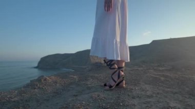 Uzun beyaz elbiseli genç sarışın bir kadın denizin kenarındaki kahverengi kayalık bir uçurumda duruyor, deniz kıyısının güzel manzarasının tadını çıkarıyor..