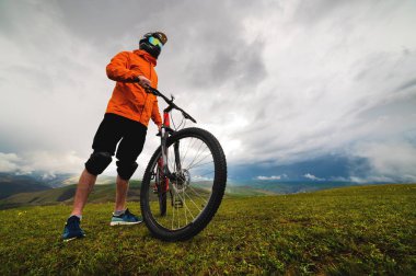 Kasklı ve korumalı bir adam bisikletle ayakta duruyor. Yeşil bir tepede, dağların arasında, yoldan çıkmanın arasında..