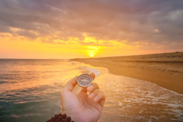 hand holds a compass against the background of the sea. the setting sun falls on the beach and ocean, there is no one on the shore.