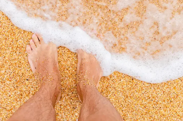 Top view of bare feet in the sand on the beach. Foaming sea texture of ocean and sand, standing barefoot on shallow sandy beach. Summer background, relaxation and vacation.