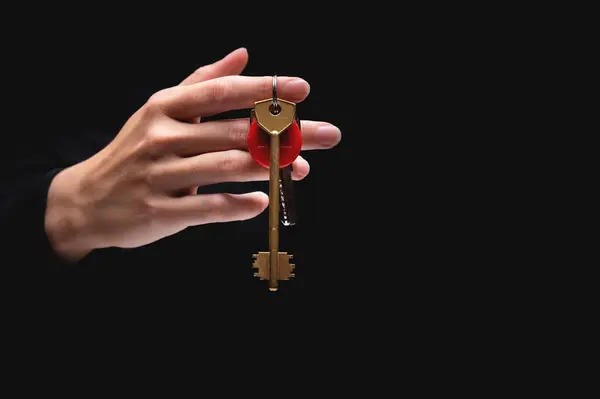 A womans hand holds keys with a round red keychain on a black background. The hand gives the keys from the darkness.