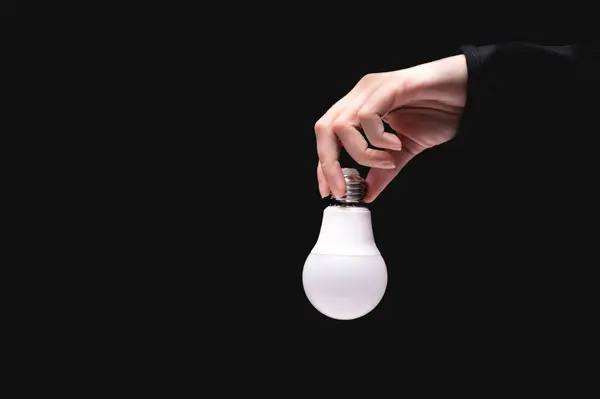 A hand holds an LED light bulb on a black background. Using an economical and environmentally friendly light bulb concept. Idea. Energy saving lamp in a womans hand