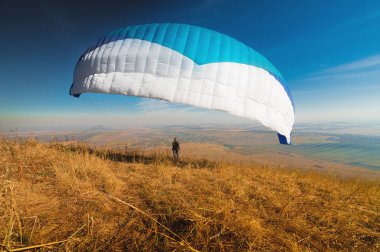 Bir paraglider, arkasında güneş ve mavi beyaz bir gölgelik olan bir dağ yamacından havalanır. Bir paraglider bir siluettir. Planör keskindir, kanat hareketi azdır. Bir erkek paraşütçü...