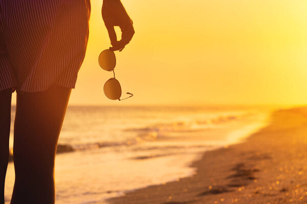 A woman in shorts stands on the shore with sunglasses in her hand, close-up. Warm colors, beach holiday at sunset.