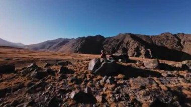 FPV gözlük ve kask takan beyaz bir adam dağlarda oturur ve etrafında uçan bir FPV akrodron kuadkopterini kontrol eder. Bir İHA pilotunun dinamik görüntüleri destansı dağların arka planında.
