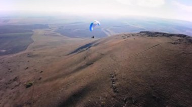 Bir FPV insansız hava aracının hava görüntüsü. Bir paraglider atleti küçük bir dağın yamaçlarına yakın uçar. Sonbaharda FPV insansız hava aracından kırsal alanlarda ücretsiz paraşüt uçuşu.