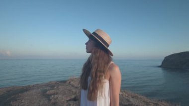 Hasır şapkalı, yüzünde gizemli bir ifadeyle dalgın bir genç kadın. Bir uçurumun kenarındaki deniz kıyısına bakar..