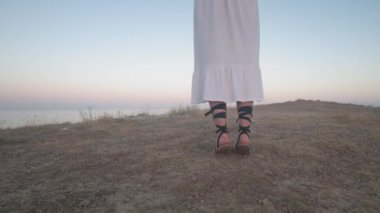 Kuru çimenlerin arasından denize doğru koşan sandaletli kadın bacakları. Beyaz elbiseli bir kızın koştuğu bir kaya..