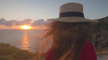 B roll - Kırmızı elbiseli ve hasır şapkalı genç bir kadın gün batımında kayalık bir deniz kıyısında duruyor. Şapkasını elinde tutuyor ve uzaklığa bakıyor. Romantik ruh hali.