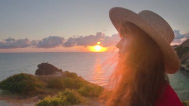 Uçurumun kenarında duran bir kız kameraya poz veriyor, gülümsüyor, rüzgar uzun saçlarıyla oynuyor. Gün batımında denizde kırmızı elbiseli genç bir kadın..