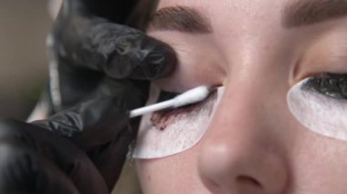 Kirpik boyama işlemi yaptıran bir kadının yakın plan portresi. Eldiven giyen bir tamirci pamuklu bezle fazla boyayı temizler..