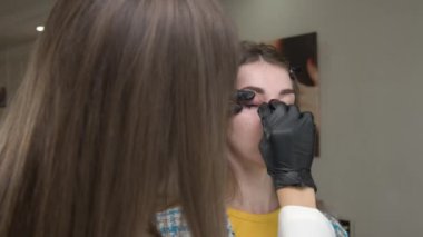 Güzellik uzmanı müşterilerin kaşlarını temizliyor. Kaşları kalıcı renge hazırlıyorum. Kafkasyalı genç bir kadının yüzüne ve bir kozmetikçinin ellerine yakından bakın.