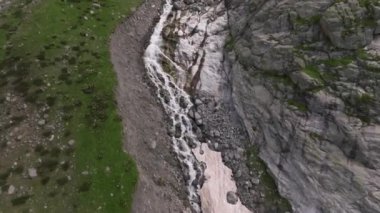 Dağlardaki bir şelalenin üzerindeki bir drondan vuruldu. Güzel manzara, hızlı akan dağ şelalesi yemyeşil çimenler ve karlı.