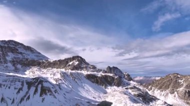 Güneşli bir havada, beyaz bulutlu, karlı bir dağ manzarası. İnsansız hava aracı, karlı dağların manzarasını, vadinin manzarasını gözler önüne seriyor..