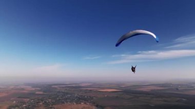 Hava görüntüsü, ufuk boyunca sabit görüntü. Etrafı tarlalar ve tepelerle çevrili bir erkek paraşütçü. Sonbaharda. Güneşli havada mavi-beyaz paraglider kanadı.