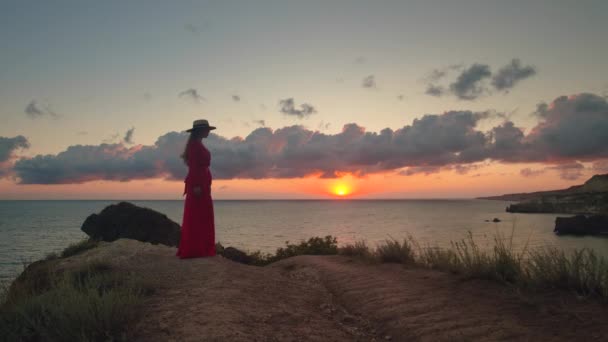 日落时 一个穿着红色衣服和草帽的年轻女子站在岩石海岸上 他手里拿着帽子 望着远方 浪漫的心情 — 图库视频影像