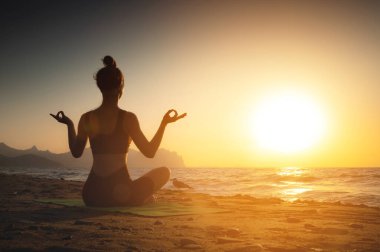 Yoga yapan kadın gün batımında meditasyon yapıyor ya da sahilde gün doğumunda. Kız lotus pozisyonunda rahatlıyor. Parmaklar çamur içinde kıvrılmış