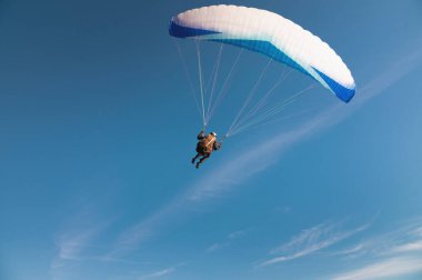 Bir paraglider, arkasında güneş ve mavi beyaz bir gölgelik olan bir dağ yamacından havalanır. Bir paraglider bir siluettir. Planör keskindir, kanat hareketi azdır. Bir erkek paraşütçü...