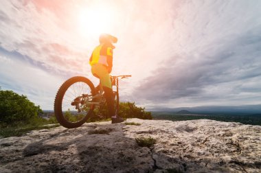 Genç bir adam dağlık bir alanda özel bir sektörün arka planına karşı bir dağ bisikletinin üzerinde oturuyor. Güneş bisikletçinin üzerinde parlıyor..