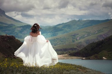 Beyaz gelinlikli bir gelin yeşil dağlarda yürüyor çimlerin üzerinde, arka planda dağlar ve bir göl var..