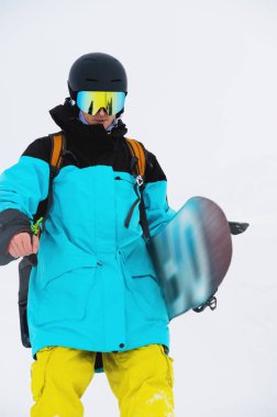 Bir erkek snowboardcunun yamaçtan aşağı doğru yürüdüğü, kask taktığı ve kar kayağıyla maske taktığı yakın plan..