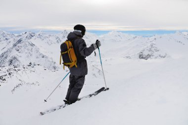 Kasklı bir atletik kayakçı ve sırt çantalı gözlükler, kayakların üzerinde, beyaz karda kayak direklerini tutarken, gökyüzüne karşı, dağların güzel manzarasının keyfini çıkarırken..