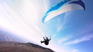 Sonbahar tepelerinin üzerinden mavi bir paraglider uçuyor, bir FPV İHA başının üzerinden ve etrafından uçuyor. Kuş bakışı.