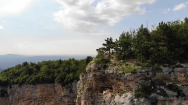 Uzun saçlı ve siyah kolsuz tişörtlü bir adam uçurumun kenarında yürüyor. Güneşli bir günde ormanla kaplı bir vadi..