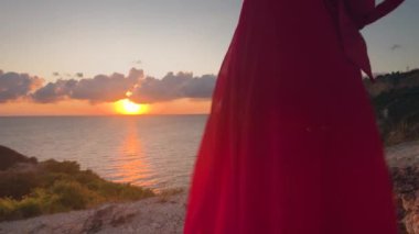 B roll - Kırmızı elbiseli ve hasır şapkalı genç bir kadın gün batımında kayalık bir deniz kıyısında duruyor. Şapkasını elinde tutuyor ve uzaklığa bakıyor. Romantik ruh hali.