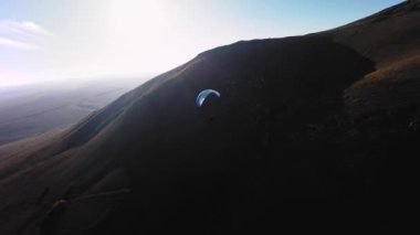 Bir FPV insansız hava aracının hava görüntüsü. Bir paraglider atleti küçük bir dağın yamaçlarına yakın uçar. Sonbaharda FPV insansız hava aracından kırsal alanlarda ücretsiz paraşüt uçuşu.