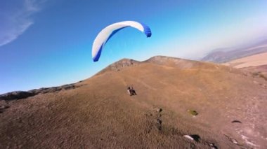 Paraşütçü bir sporcu, güneşli bir sonbahar gününde bir köyün yakınındaki küçük bir dağın yamacında yamaç paraşütü üzerinde uçar. FPV İHA kuadkopteri ile akro modda hava görüntüleme çekimi.