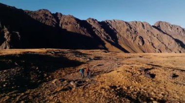 Sırt çantalı iki adam gün batımında yüksek bir plato boyunca dağlarda kar örtülü dağlara ve yüksek kayalık duvarlara karşı yürür. Hava görünümü.