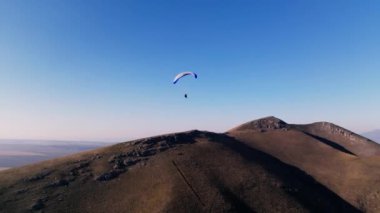 Paraglider tepelerin üzerinden uçuyor, İHA arkasından uçuyor, havada pürüzsüz hareketler yapıyor..