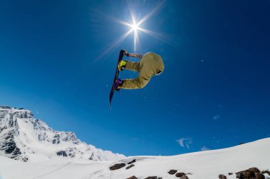 Bir snowboardcu büyük bir havadan büyük bir tahta tutarak atlar, bir burun ya da kuyruk numarası sırayla, kar kaplı dağların arka planına ve parlak güneşe karşı.