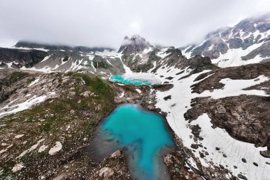Dağlarda yüksek bir alp gölünün havadan geniş açılı görüntüsü. Dağ zirveleriyle çevrili göl kısmen karla kaplı..