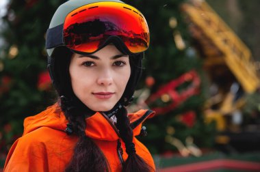 Kayak gözlüklü ve kasklı gülümseyen kadın duvara yaslanmış kameraya bakıyor, mutlu bir kızın portresi..