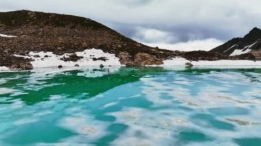 Bir adam, kısmen karla kaplı, karla kaplı bir dağ turkuaz gölündeki bir kürek tahtasının üzerinde yüzüyor. Hava görünümü.