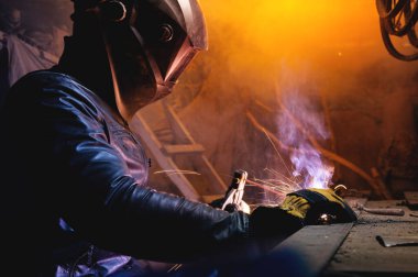 Deri ceketli bir kaynakçı ve kaynak maskesiyle eski bir ahırın karanlık bir odasında kaynak yapıyor. Ev kaynaklama.