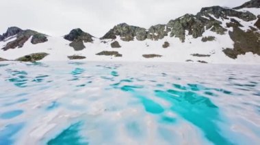 FPV insansız hava aracı bulutlu bir gölün üzerinde uçuyor ve arka planda yüksek kayalık dağlar olan bir dağ sırası var..