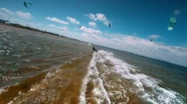 Güneşli bir yaz gününde dalgıç kıyafetiyle denizde dalgalara binen zinde bir uçurtmacının arka ve yan görüntüsü. İnsansız hava aracı atletin arkasından uçuyor, yavaş çekim..