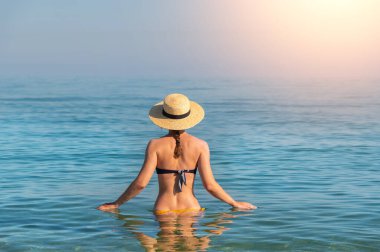 Farklı bir mayo ve hasır şapkalı beyaz bir kadın güneşli bir günde beline kadar deniz suyuna batmış durumda. Sırtı kameraya dönük..