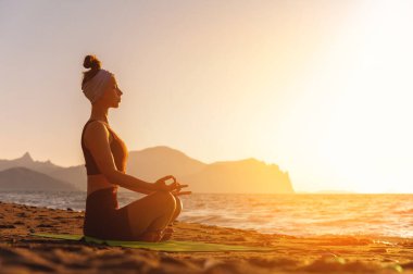Lotus poz veren genç bir kadın okyanus kıyısında sağlıklı bir yaşam için yoga yapıyor. Kadın gücünü ve ruhunu geri kazanmak için egzersiz yapıyor. Yoga pozu deniz kıyısında gün batımında.