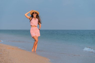 Yaz kumsalı tatili konsepti, şapkalı ve plaj kıyafetli mutlu genç kadın ellerini havaya kaldırıp sıcak yaz akşamında plaj ve okyanus manzarasının tadını çıkarıyor..