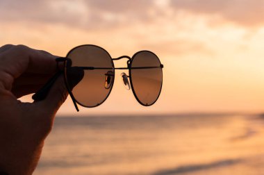 Güneş gözlüğü tutan bir kadının eli denizin ve gün batımının arka planına yakın çekim..