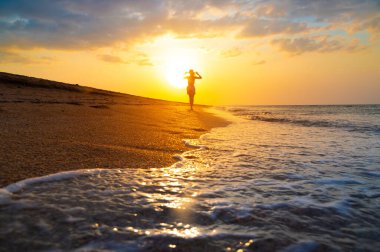 Gün batımında deniz kıyısında ya da okyanusta yürüyen şapkalı ince beyaz bir kadın. Düşük açılı su geniş açılı.